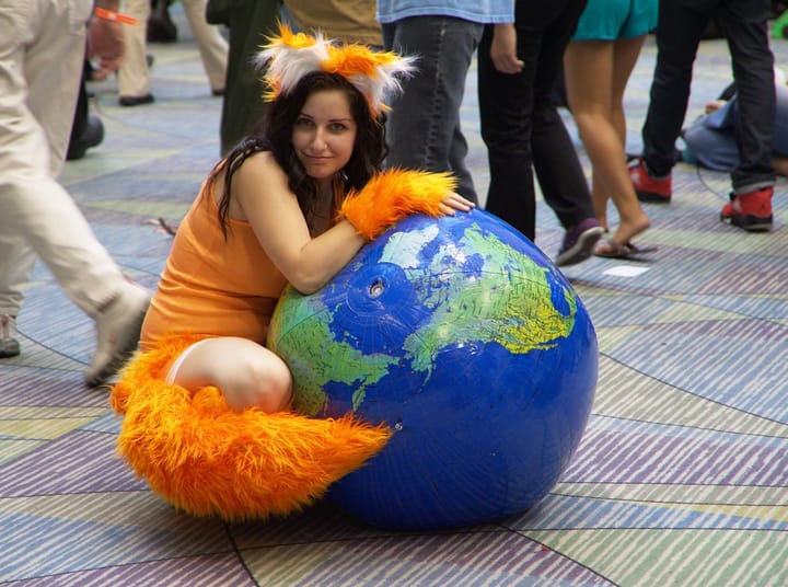 A woman in an orange-eared and cuffed Firefox cosplay hugging an inflatable globe, smiling as she sits on a patterned floor.